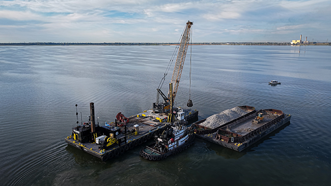 An aerial photo of a tug pushing a barge loaded with cobble. An empty barge with a front-end loader sits to the right side. On the left side is a third barge with a tall crane with attached grapple and other construction equipment. The tug and barges are surrounded by calm water and land is visible on the horizon. The tug and barges look to be within a few miles from shore.